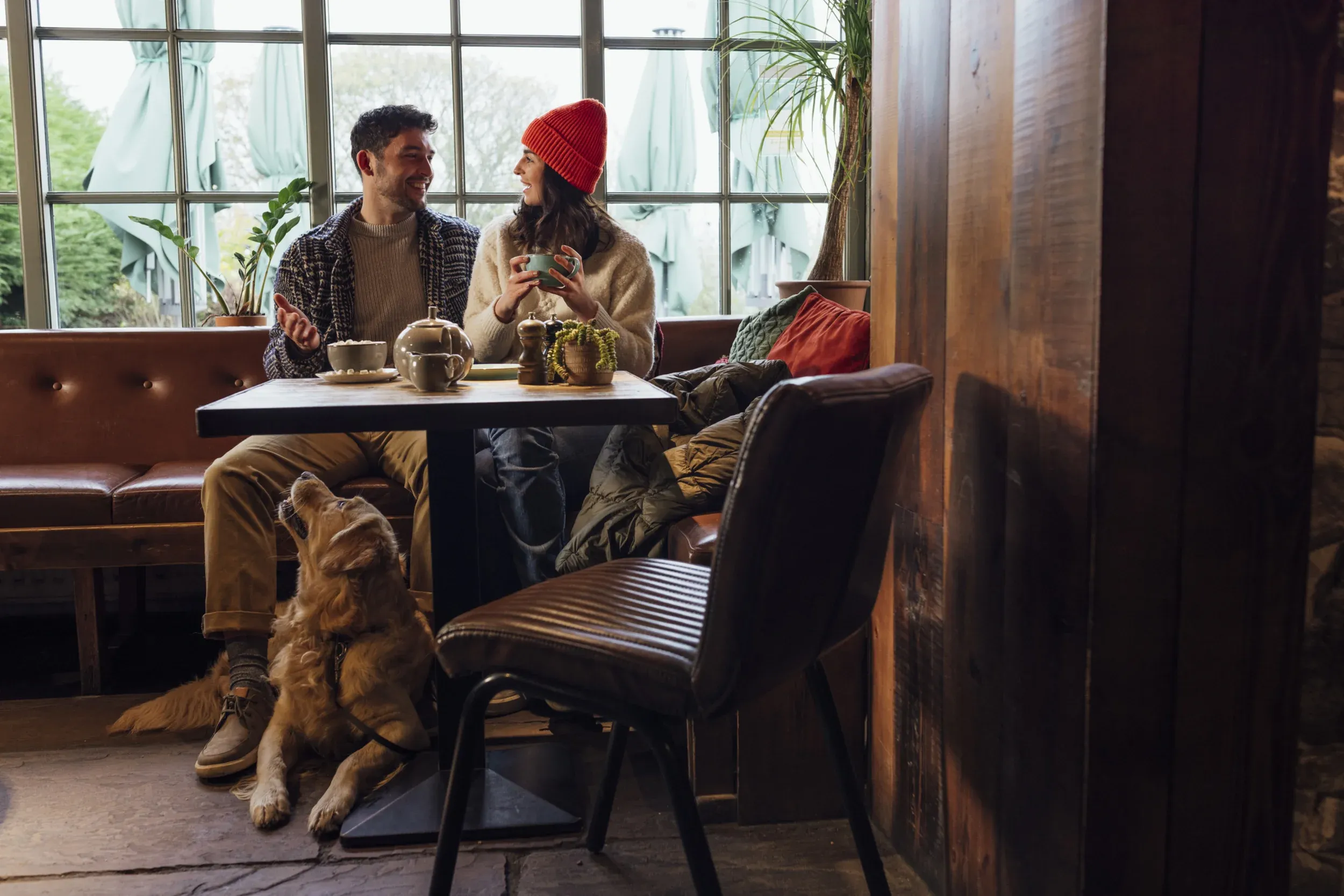 young man and woman sitting at a table in a cozy, rustic coffee shop, smiling as they talk and are wearing warm clothing. a golden retriever on a leash is laying at their feet and looking up at them and thinking "please pet me".
